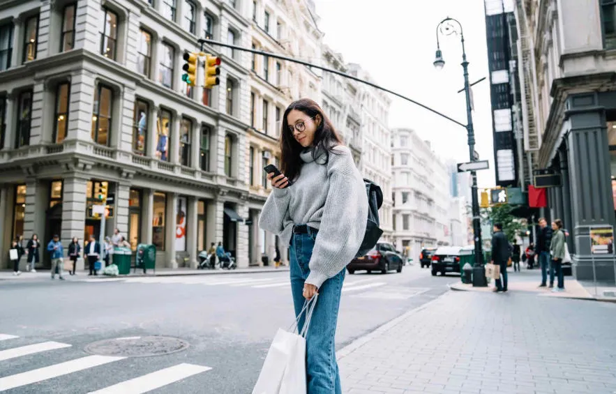 A young woman stands at a city crosswalk checking her phone, holding shopping bags, surrounded by historic buildings and light traffic.