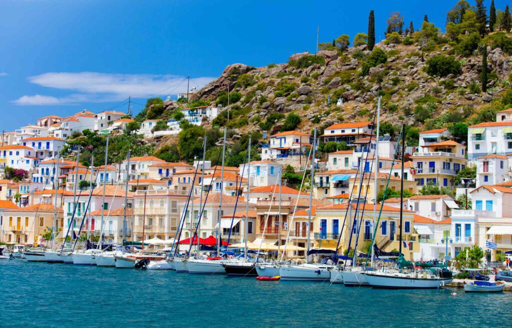Colorful coastal village in Greece with sailboats lined along the waterfront and homes built into a rocky hillside.