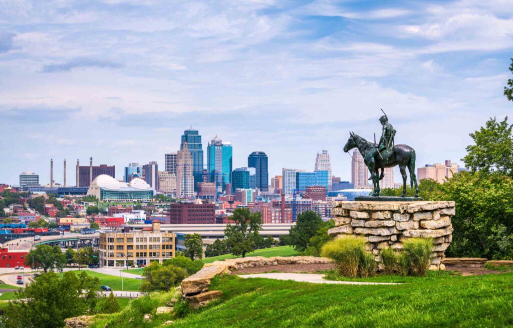 Panoramic view of downtown Kansas City, Missouri with the Scout statue in the foreground overlooking modern skyscrapers under a partly cloudy sky.