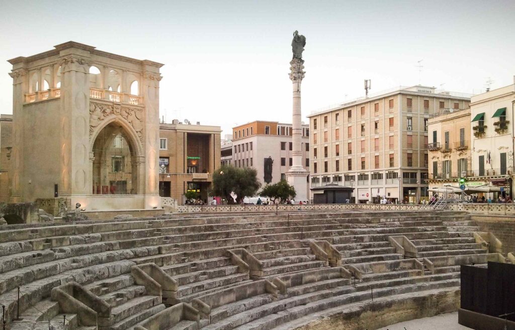 An ancient Roman amphitheater in Lecce, Italy, surrounded by historic buildings and a decorative stone arch.