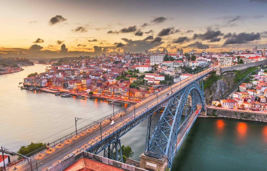 A panoramic view of Porto, Portugal, featuring the Dom Luís I Bridge spanning the Douro River at sunset.