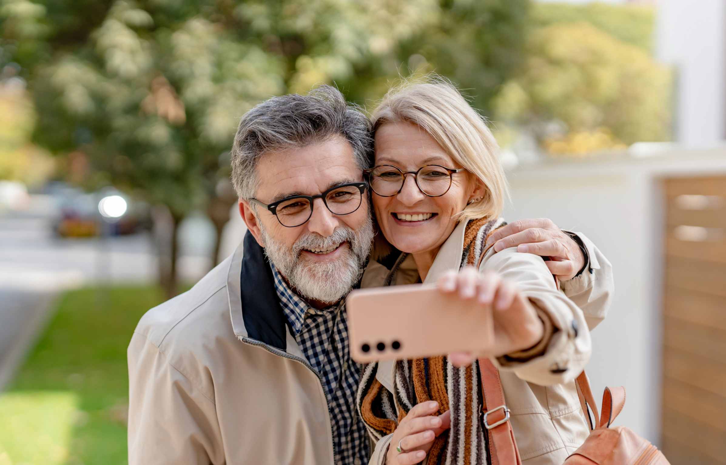 Smiling retired couple taking a selfie outdoors during a peaceful walk, wearing glasses and light jackets.