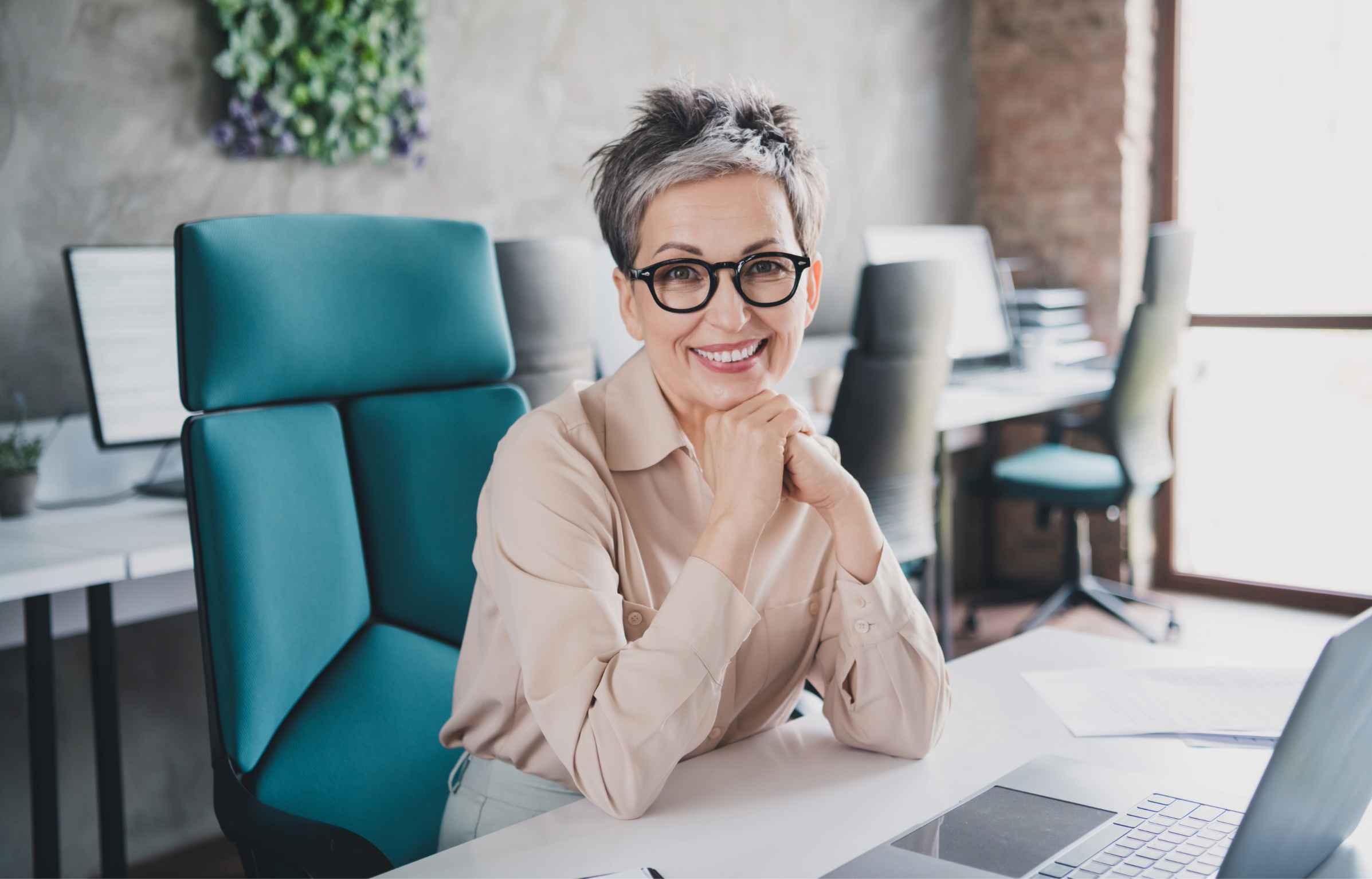 A senior professional woman sitting at a desk in a modern office, smiling confidently with her hands resting under her chin, with a laptop and workspace around her.