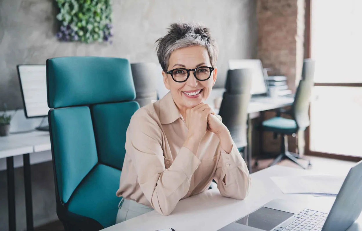 A senior professional woman sitting at a desk in a modern office, smiling confidently with her hands resting under her chin, with a laptop and workspace around her.