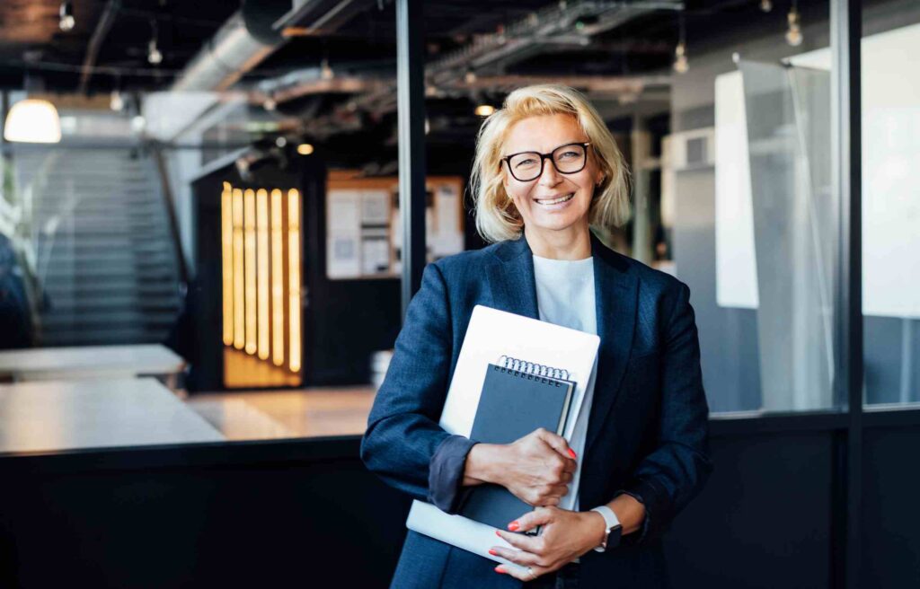 A confident senior businesswoman standing in an office environment, holding a notebook and laptop, and smiling warmly at the camera while representing relocating senior executives.