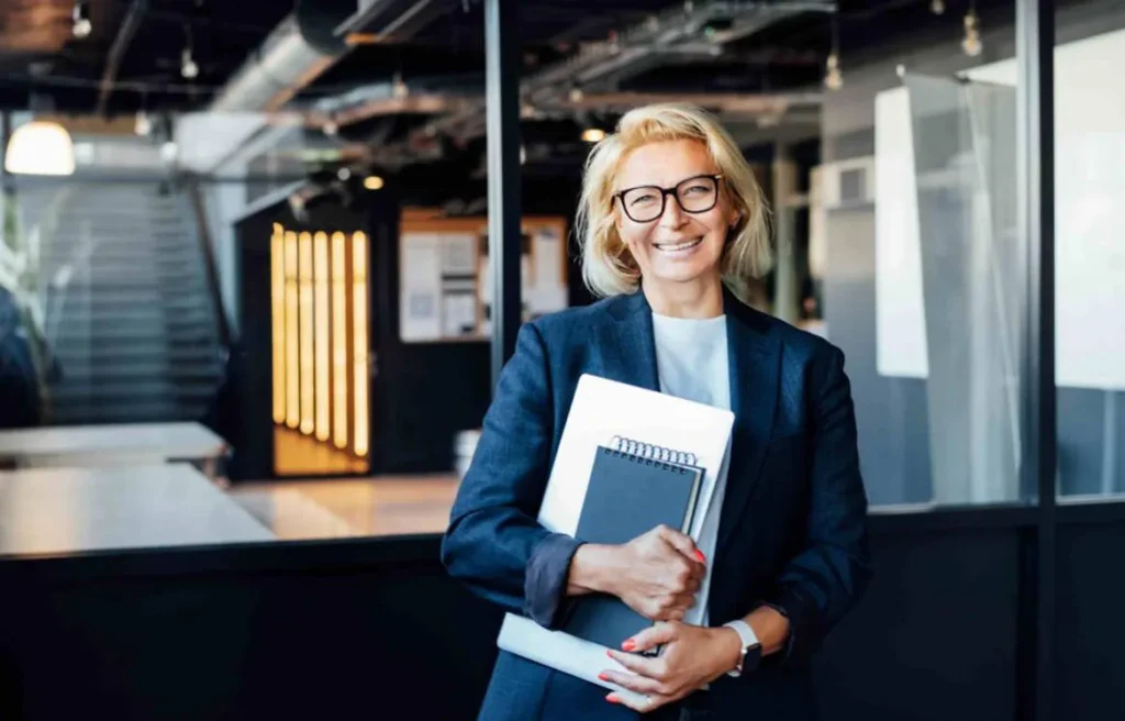 A confident senior businesswoman standing in an office environment, holding a notebook and laptop, and smiling warmly at the camera while representing relocating senior executives.
