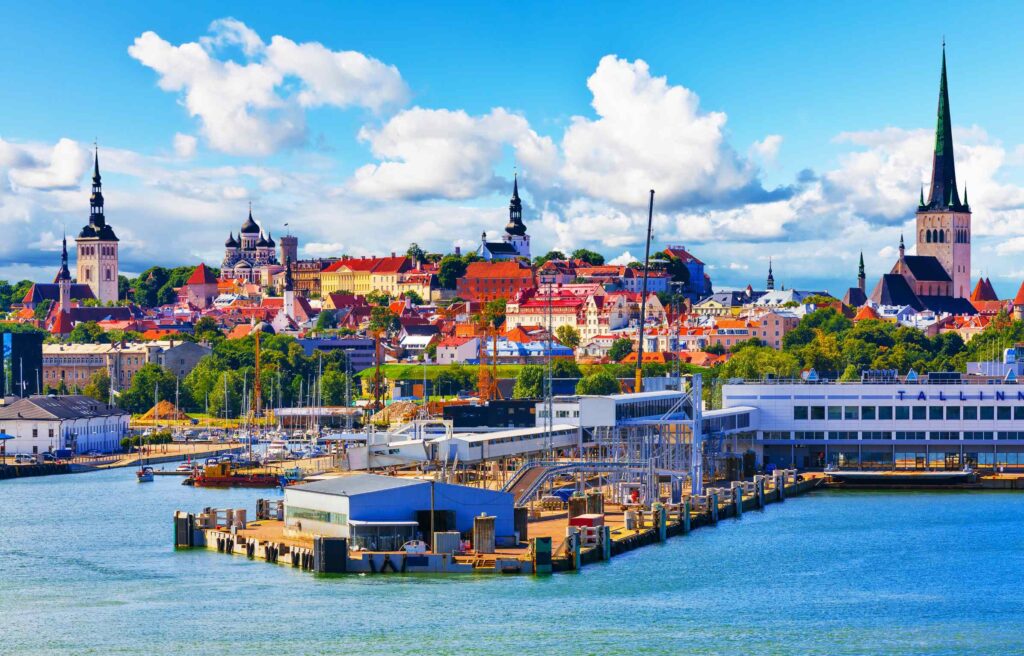 A panoramic view of Tallinn, Estonia, showing the harbor and the colorful medieval old town with church spires under a bright sky.