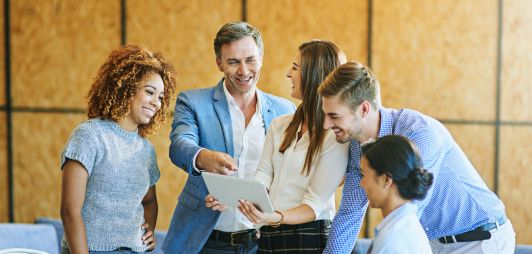 A diverse group of professionals gathered around a table in a modern office, smiling and discussing ideas while reviewing information on a digital tablet during a team meeting.