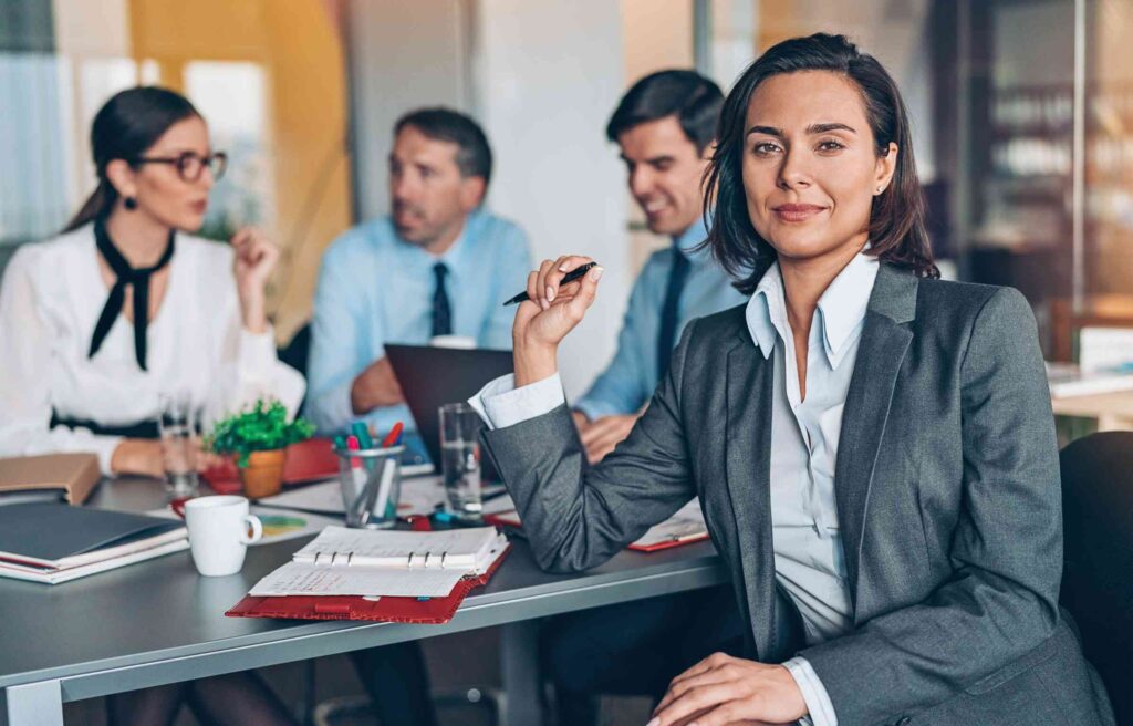 A businesswoman in a gray suit sits confidently at a meeting table while her team reviews tech team relocation details in the background.