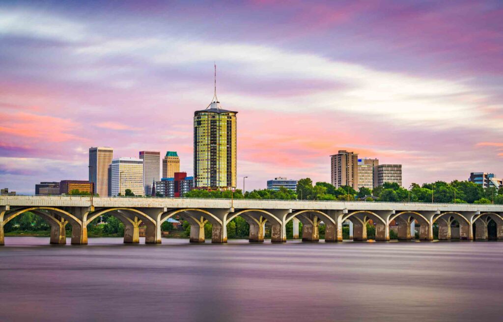 Tulsa, Oklahoma skyline at sunset with colorful clouds, a bridge over the Arkansas River, and modern high-rise buildings.