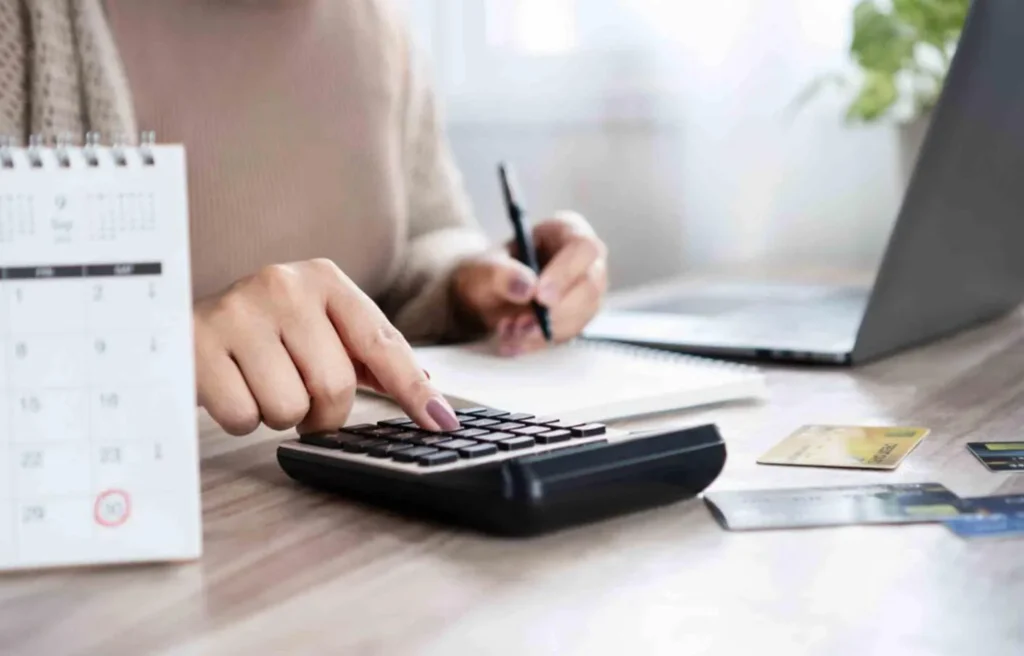 A woman using a calculator and taking notes beside credit cards and a calendar, representing budgeting and planning for relocating to “low-cost cities”.