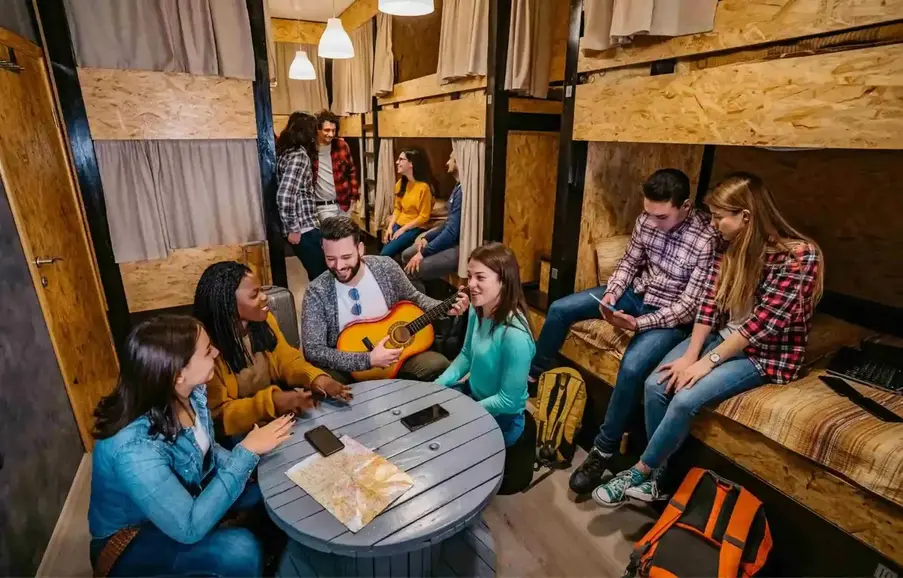 Hostelworld hostel common room with travelers talking playing guitar and socializing in shared bunk space.