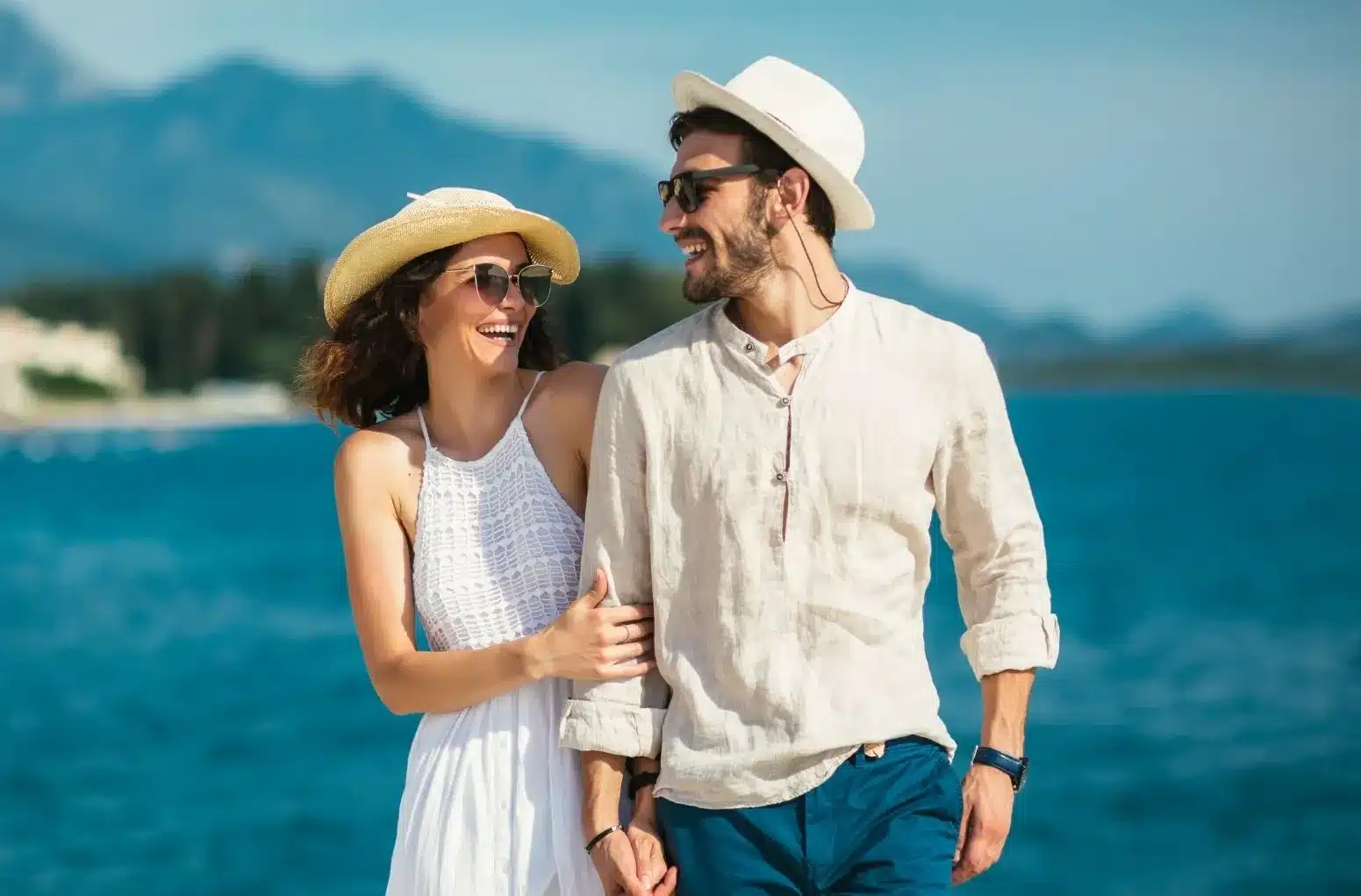 Joyful couple walking hand in hand along the waterfront, dressed in summer outfits and smiling warmly