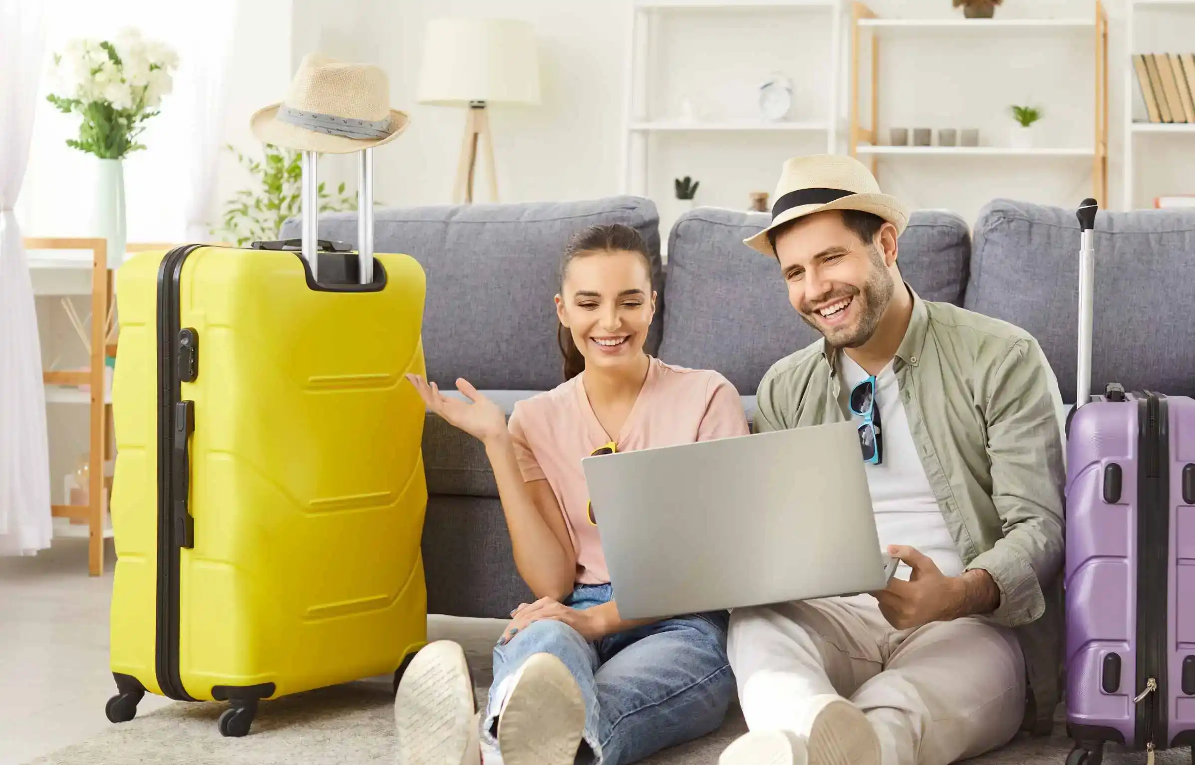 Couple planning a trip online with a laptop at home, sitting beside packed suitcases ready for travel