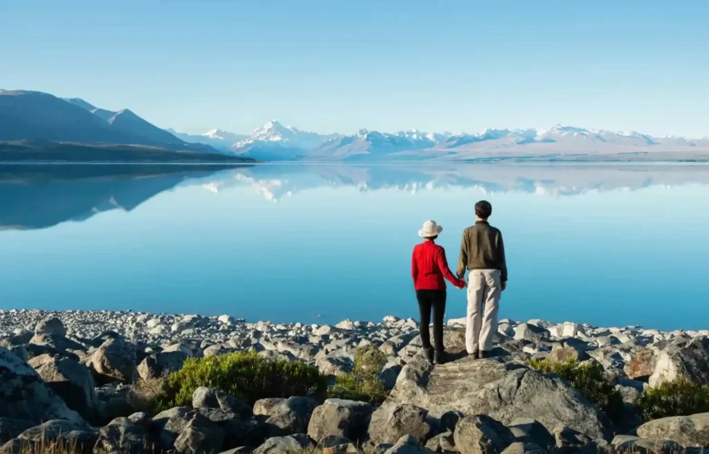 A couple standing on rocky ground overlooking a calm blue lake with snow capped mountains reflected in the water in New Zealand.