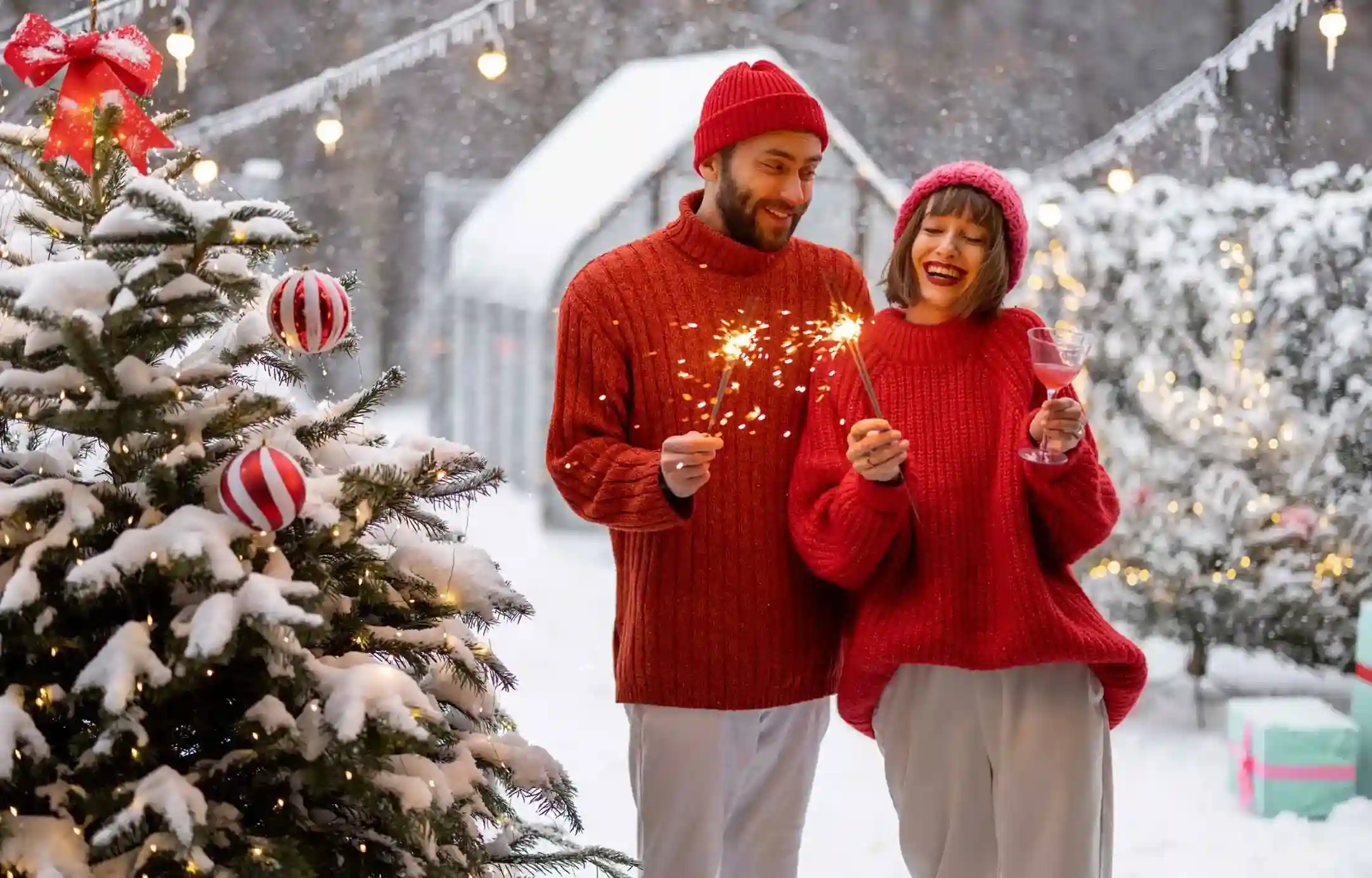 Couple in red sweaters celebrating beside a snow-covered Christmas tree, enjoying winter festivities in one of the best places to spend Christmas.