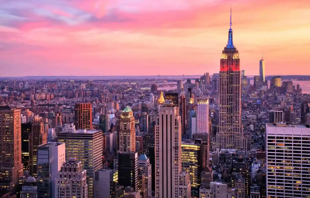 New York City skyline at sunset with the Empire State Building lit in holiday colors and dense city lights below.