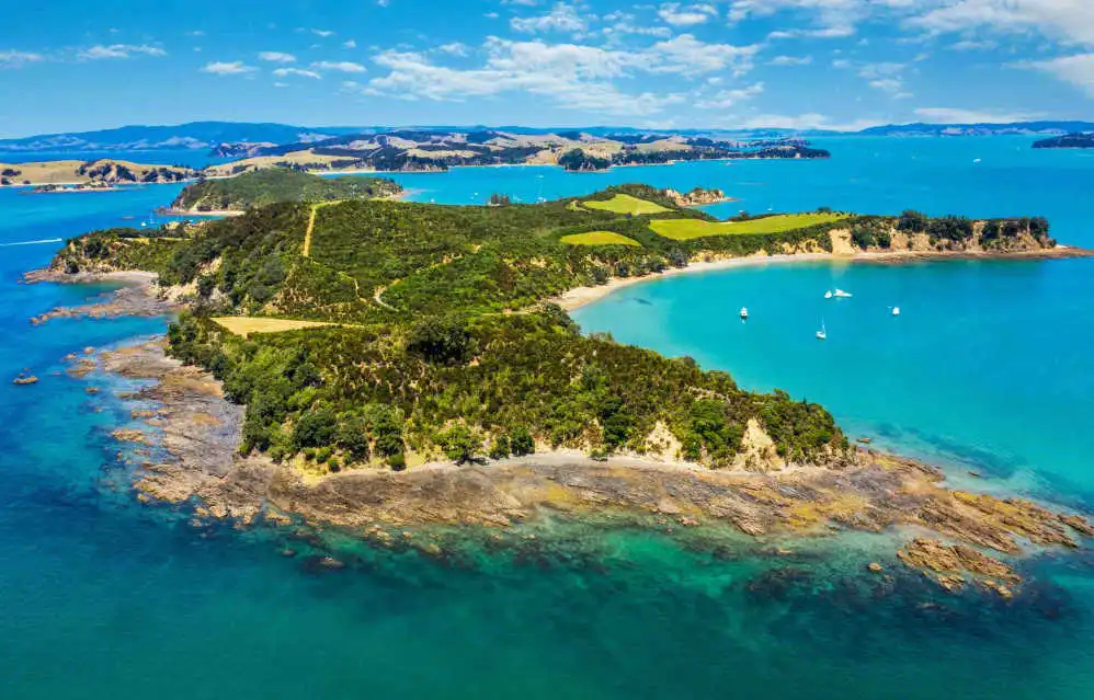 An aerial view of a green coastal landscape in New Zealand with blue ocean water, small boats anchored in coves, and rolling hills in the distance, reflecting the appeal of new zealand investor visa citizenship. 
