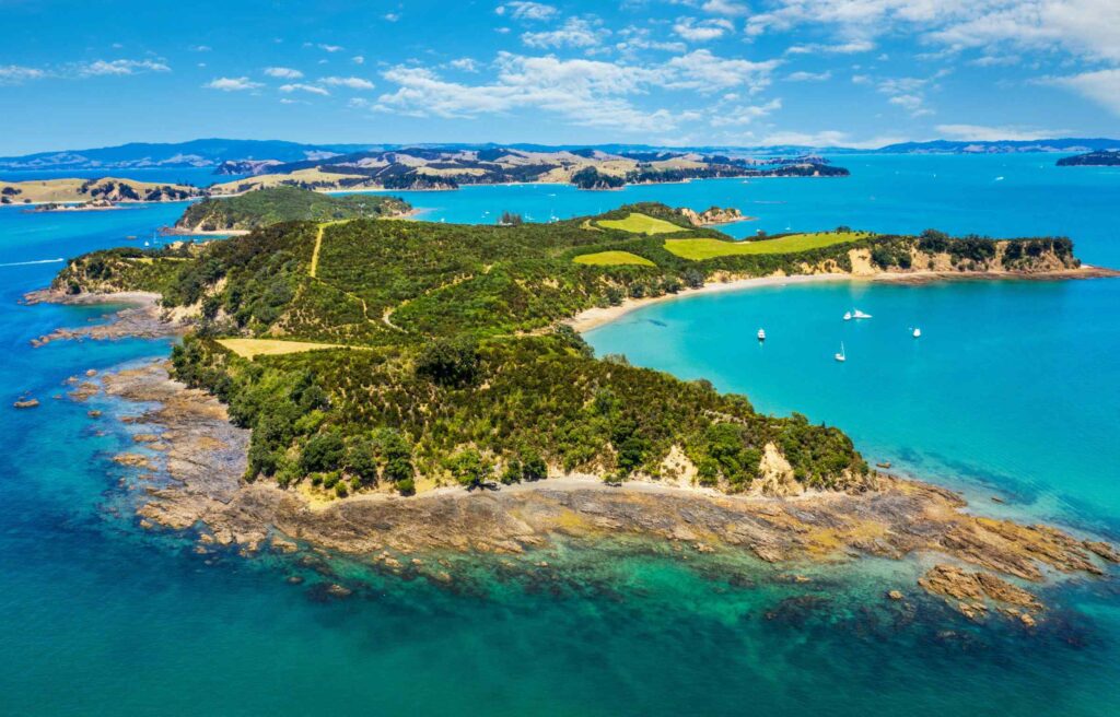 An aerial view of a green coastal landscape in New Zealand with blue ocean water, small boats anchored in coves, and rolling hills in the distance, reflecting the appeal of new zealand investor visa citizenship.