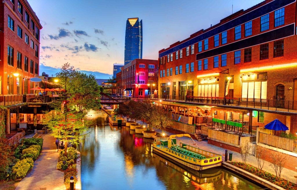 Scenic view of Oklahoma City’s Bricktown Canal at dusk with lit restaurants and boats.