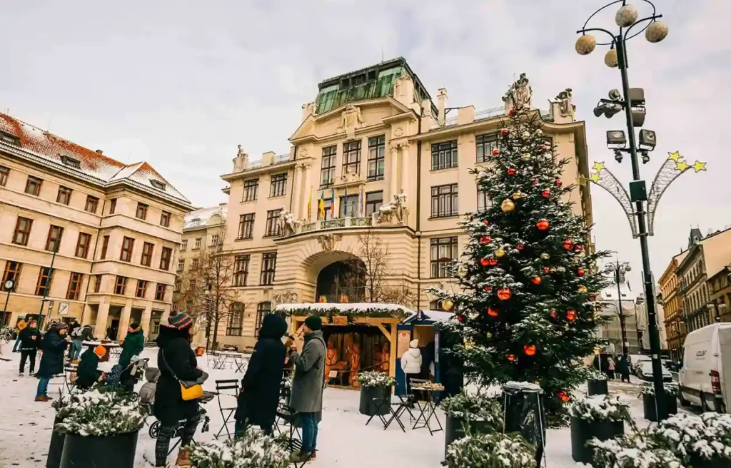Prague Old Town Square in winter with a decorated Christmas tree, market stalls, historic buildings, and light snowfall.