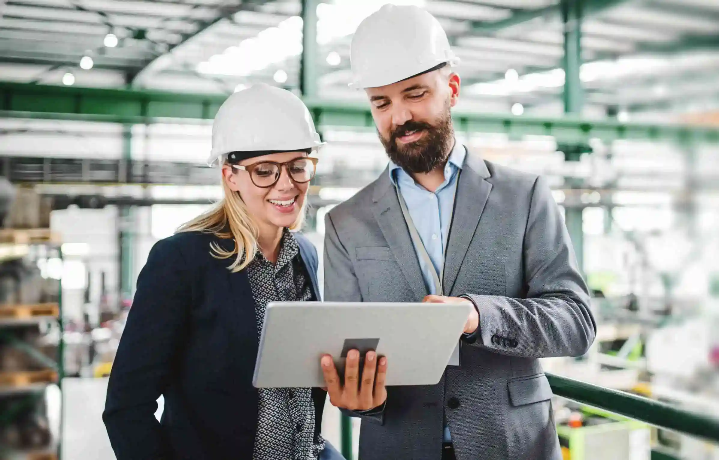 Two engineers wearing hard hats review project data together on a tablet inside an industrial facility.