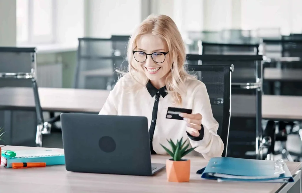 Smiling woman using a laptop and holding a credit card at an office desk while shopping online.