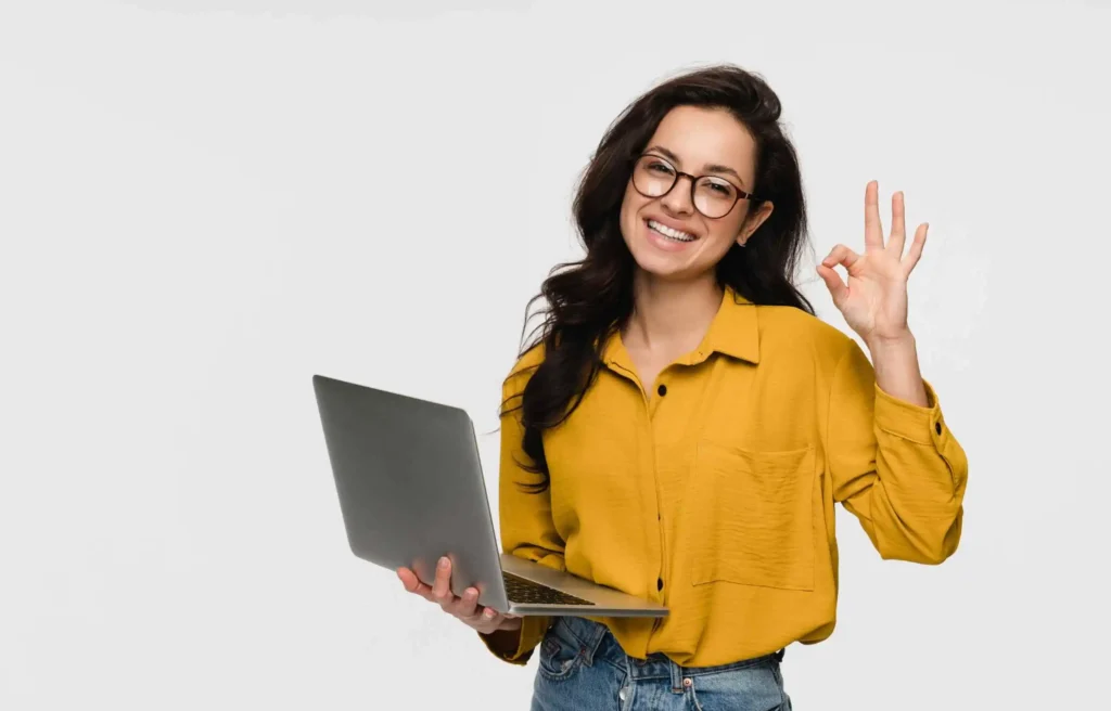 Professional woman holding a laptop, smiling and making an OK hand gesture on a light background.