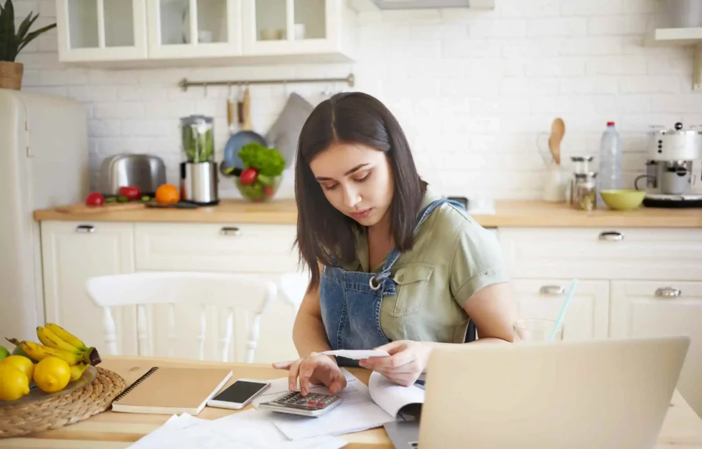 Woman paying bills and reviewing receipts with calculator and laptop in a home kitchen.