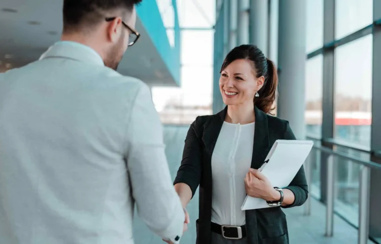 Businesswoman shaking hands with male professional in modern office hallway holding documents.