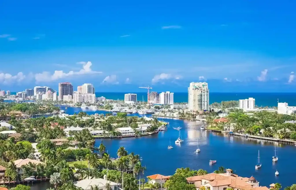 Florida coastal skyline with waterfront homes, yachts, palm trees, and high-rise buildings under a bright blue sky.
