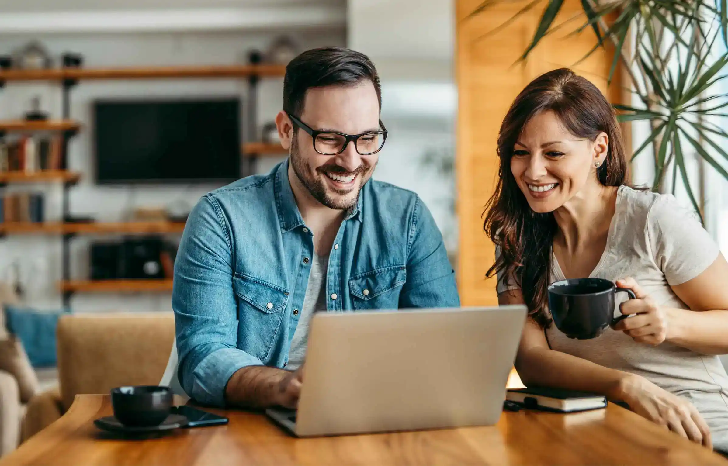 Happy couple smiling at laptop while discussing retirement plan and future financial goals at home.