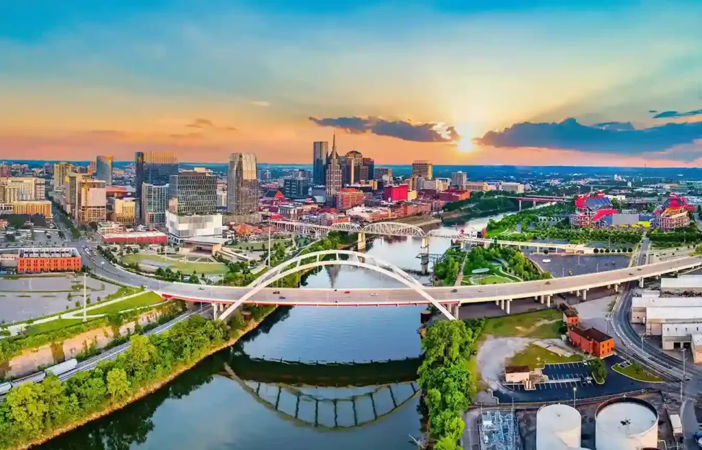 Nashville downtown skyline at sunset with river bridges, modern buildings, and city lights reflecting on the water.