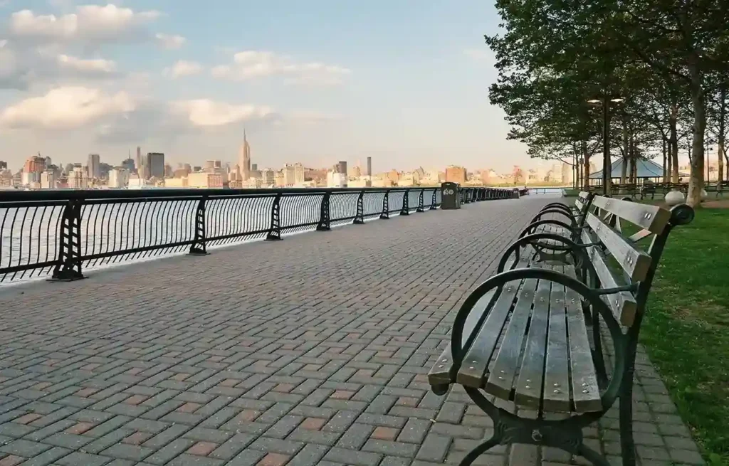New Jersey waterfront promenade with benches and Manhattan skyline visible across the Hudson River.