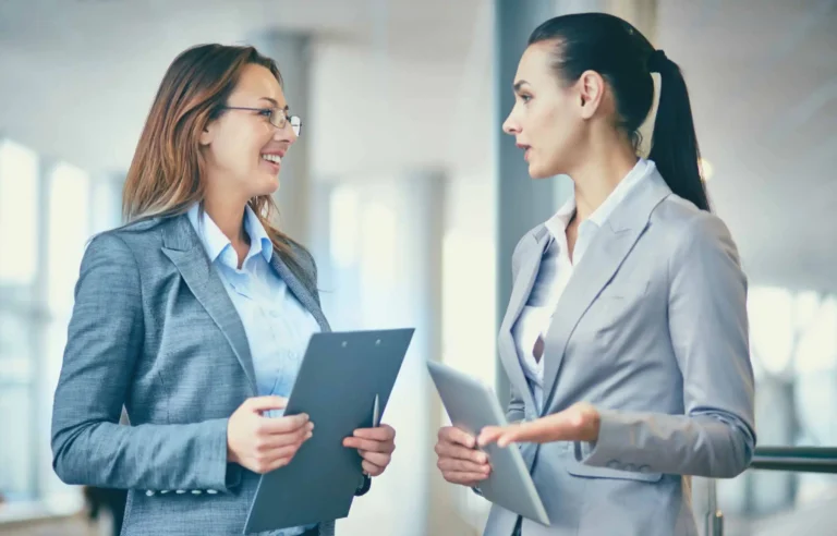 Professional women discussing relocation documents and employee mobility plans in a modern office corridor.