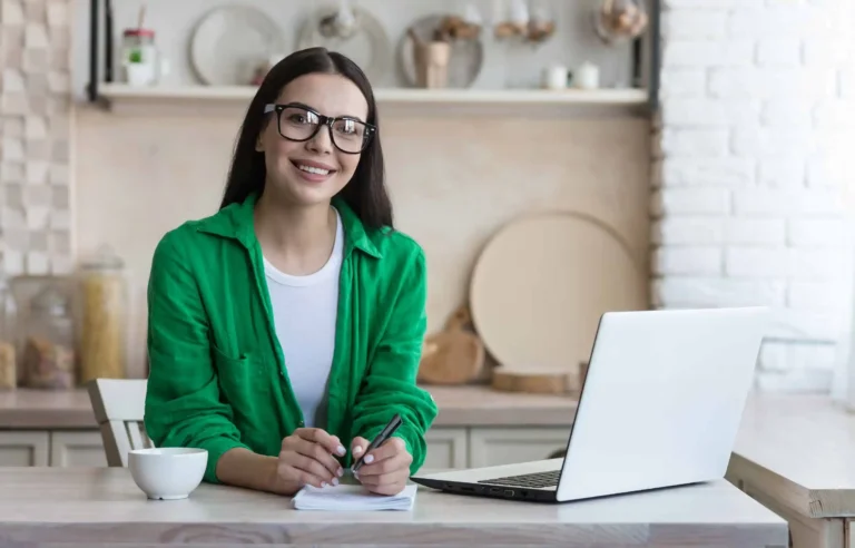 Remote work woman with glasses writing notes beside laptop at kitchen workspace.