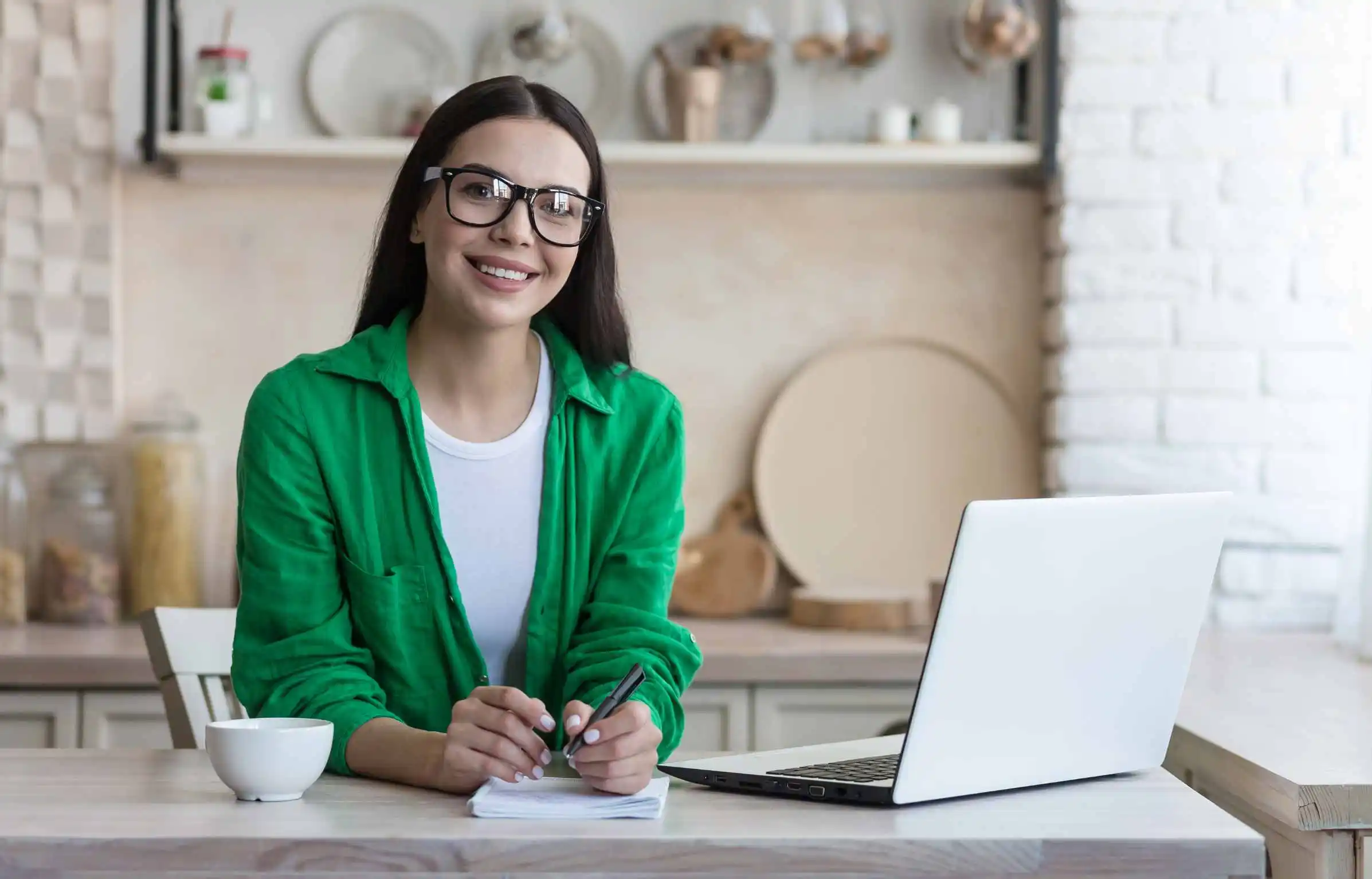 Remote work woman with glasses writing notes beside laptop at kitchen workspace.