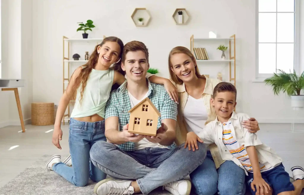 Smiling family sitting on floor holding small house model symbolizing relocating with family and new home planning.