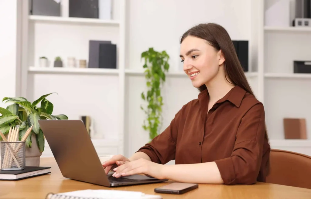 Smiling woman working on laptop at home desk representing most in-demand remote jobs with plants and phone nearby.