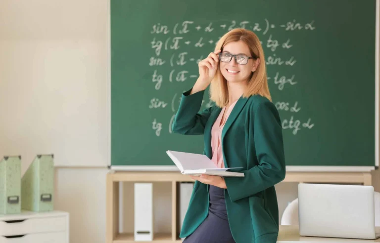 Teacher holding a notebook and adjusting glasses while standing in front of a classroom chalkboard with math formulas.