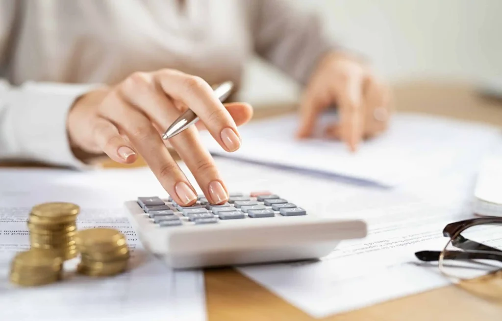 Woman using calculator to estimate corporate relocation management costs with documents and coins on desk.