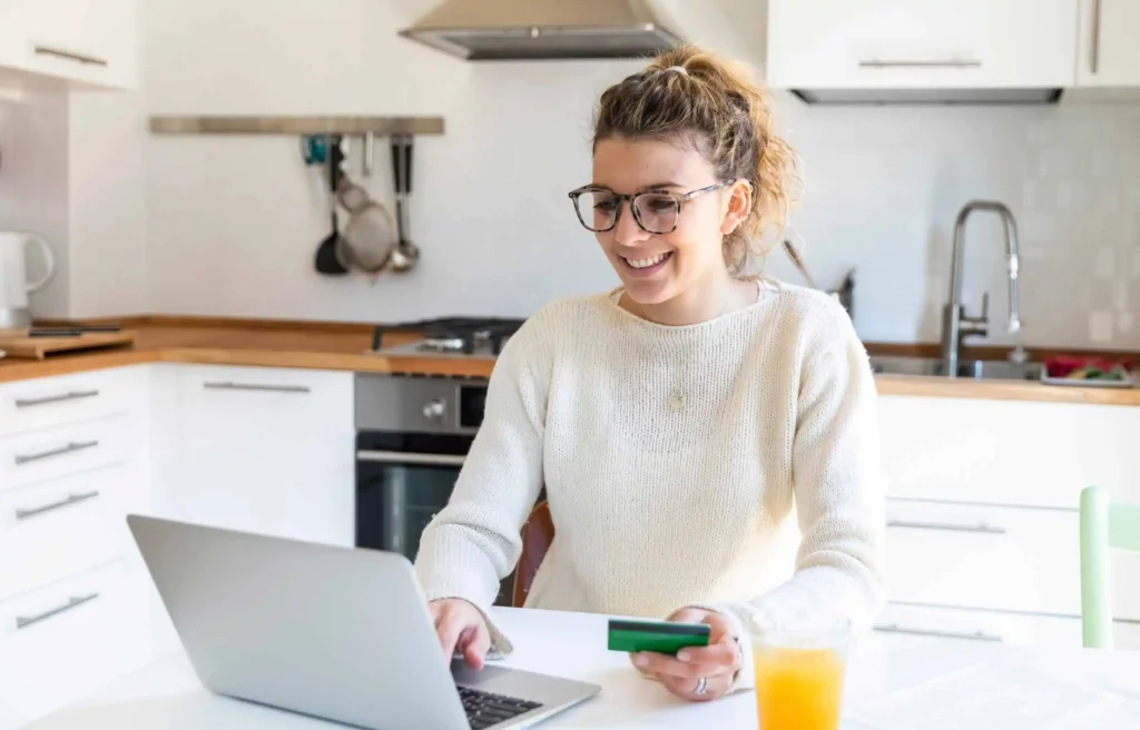 A remote worker using a laptop and credit cards to fund a move during online payment in the home kitchen.