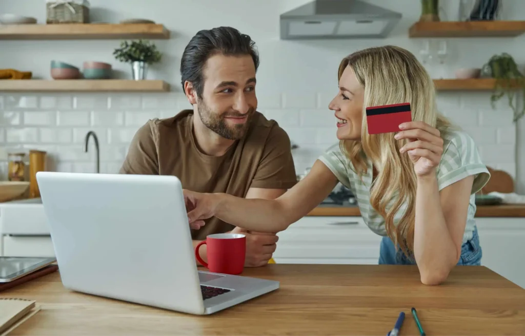 A woman showing a credit card to her partner while browsing on a laptop at home.
