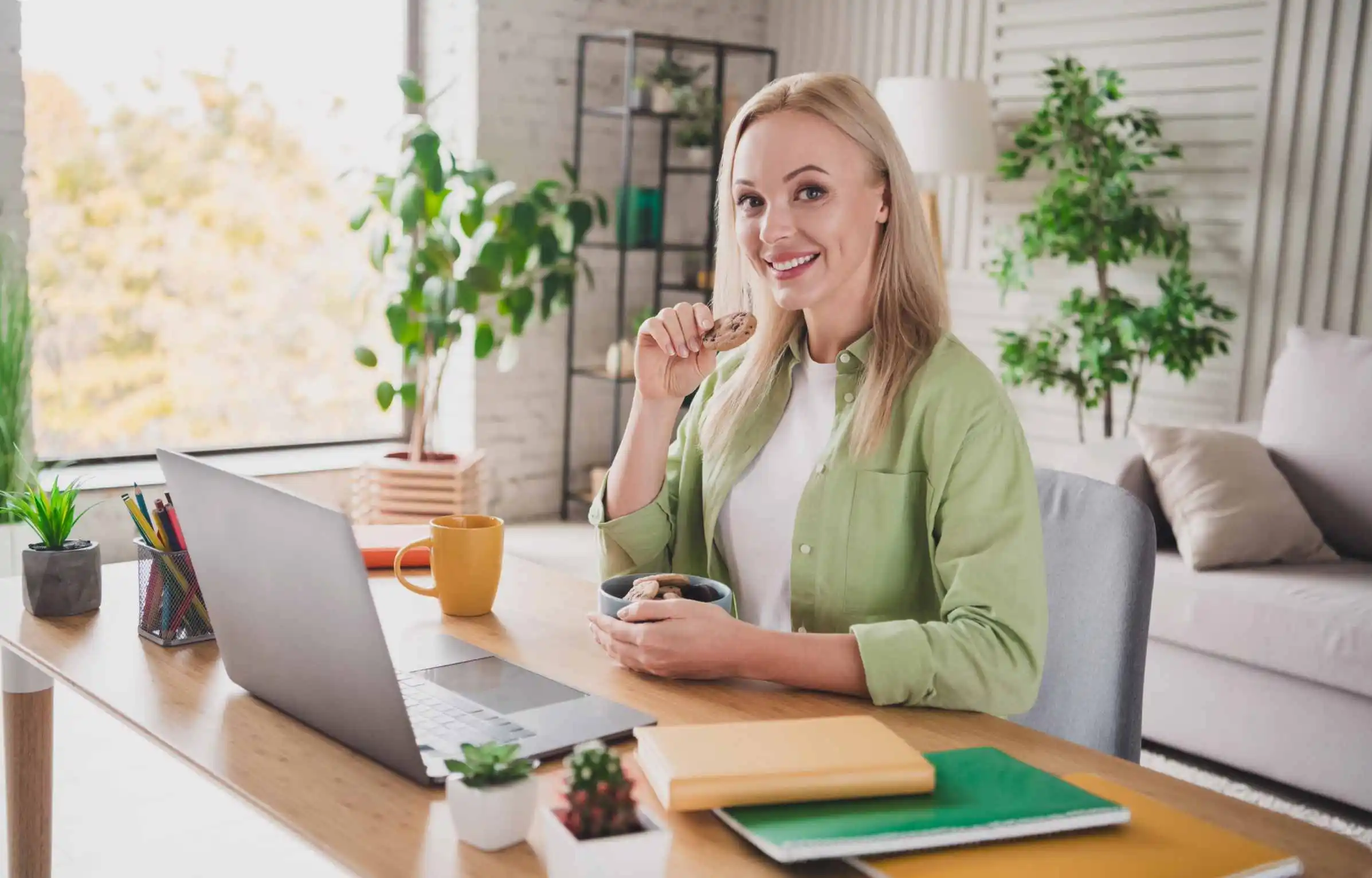 Woman working remotely at desk with laptop and snack in a bright home office.