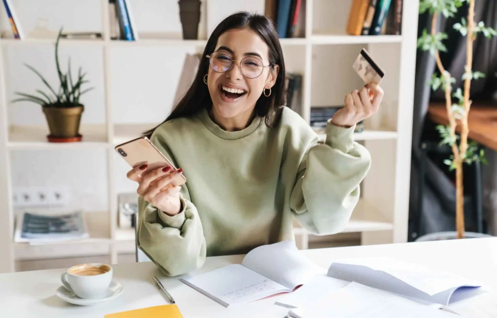 Student smiling at a desk while holding a phone and a card.
