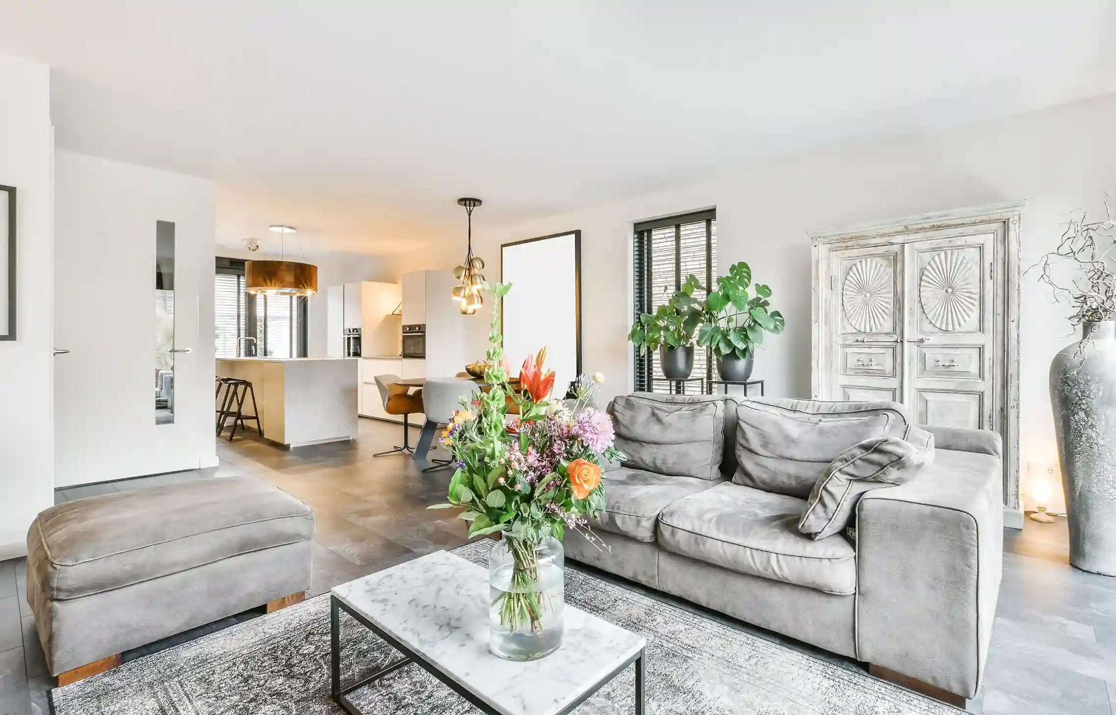 Bright furnished corporate apartment with a gray sofa, modern kitchen, plants, and fresh flowers.