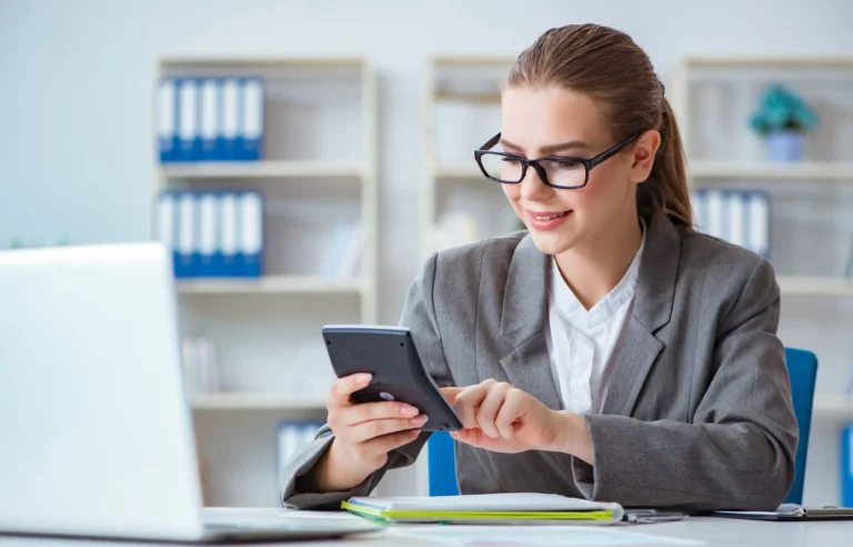 Businesswoman using a calculator at her desk beside a laptop and paperwork.