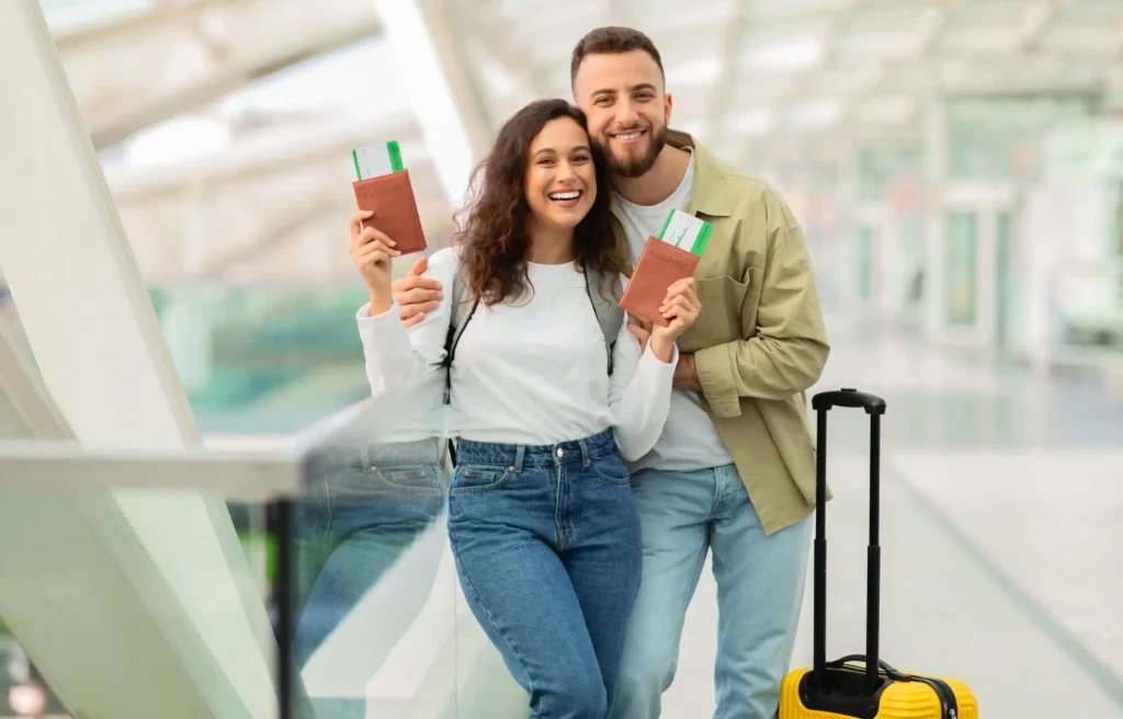 Couple at an airport holding passports and boarding passes while planning moving expenses beside a yellow suitcase.