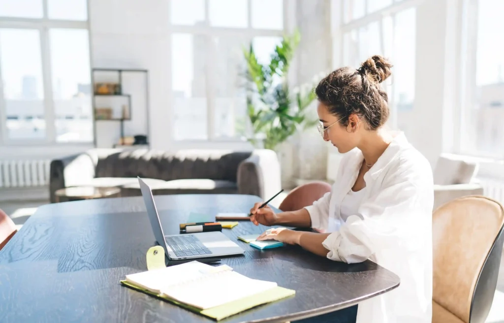 Relocating professional working on a laptop at a dining table in a bright, furnished apartment, corporate housing vs. housing stipend.