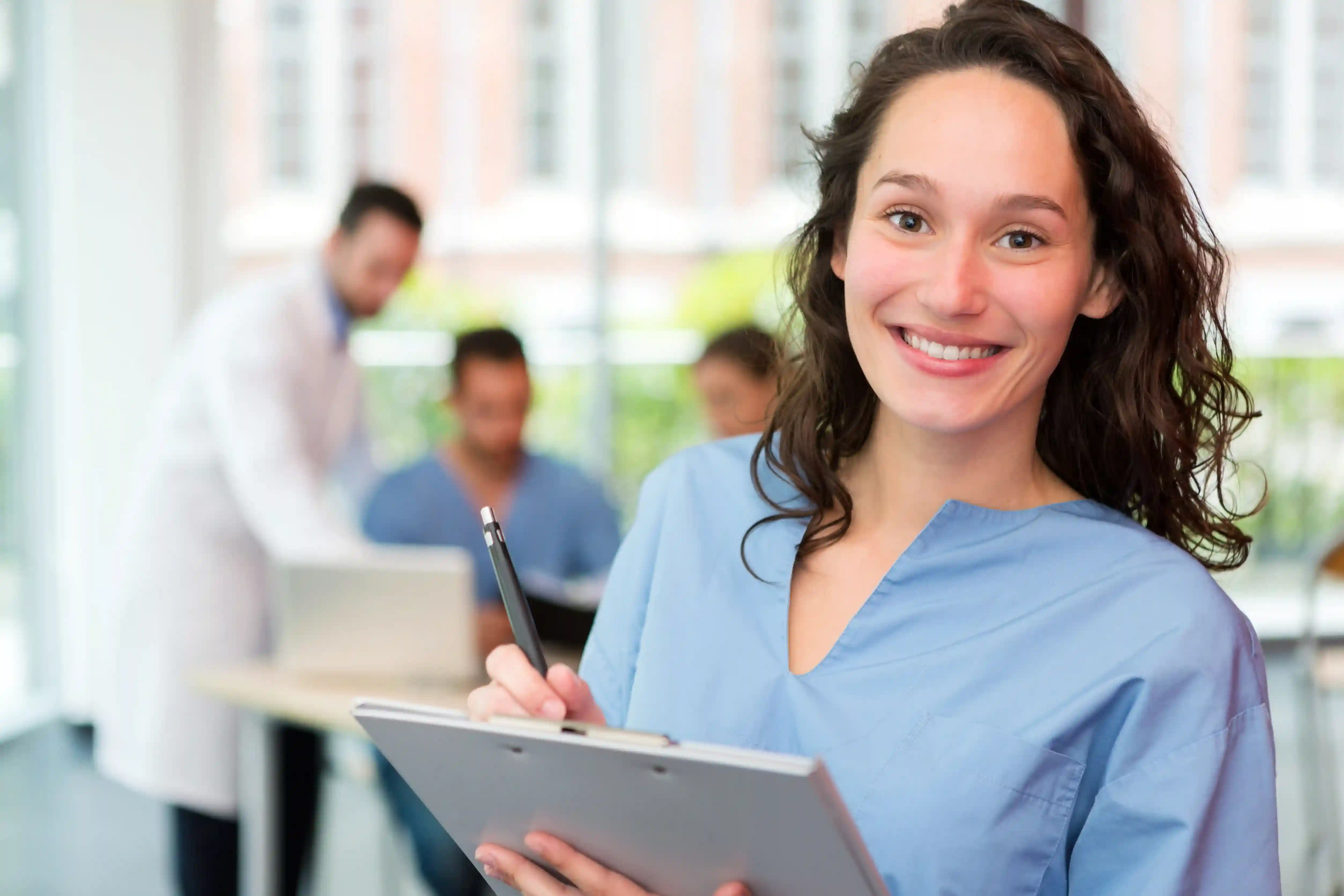 Smiling nurse in blue scrubs holding a clipboard at a hospital while preparing for a nursing relocation.
